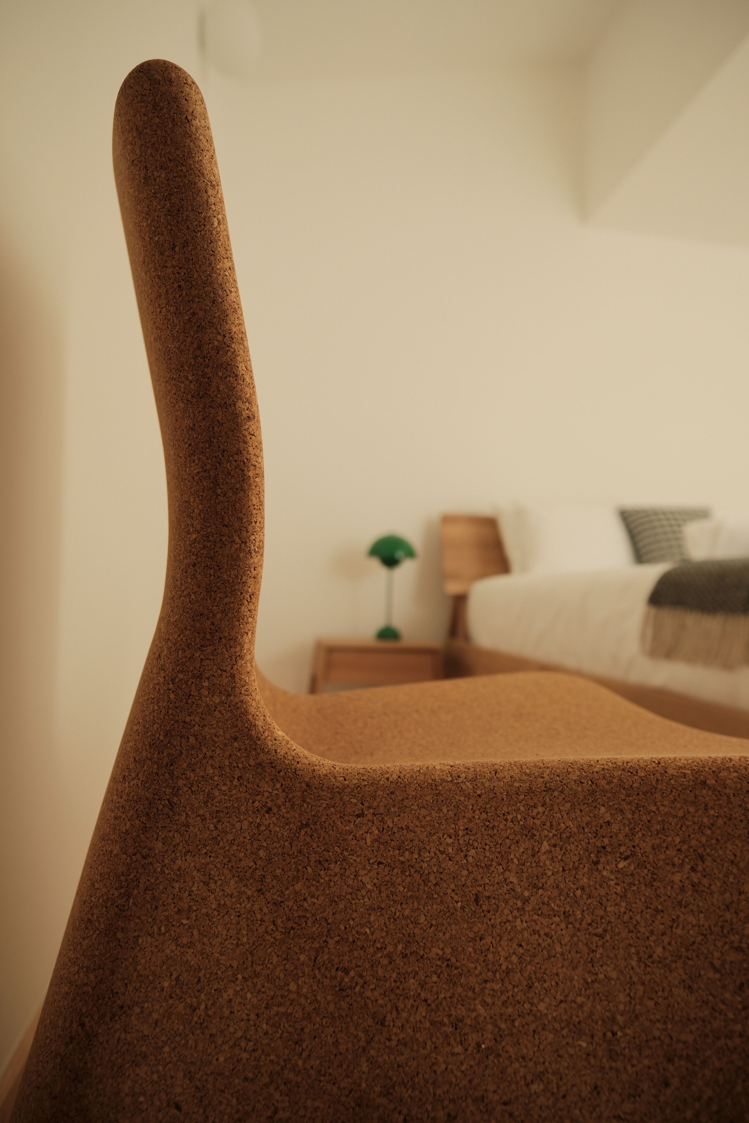 A close-up of a textured, brown chair with a curved backrest. In the background, there is a bed with a white duvet and a green lamp on a bedside table.