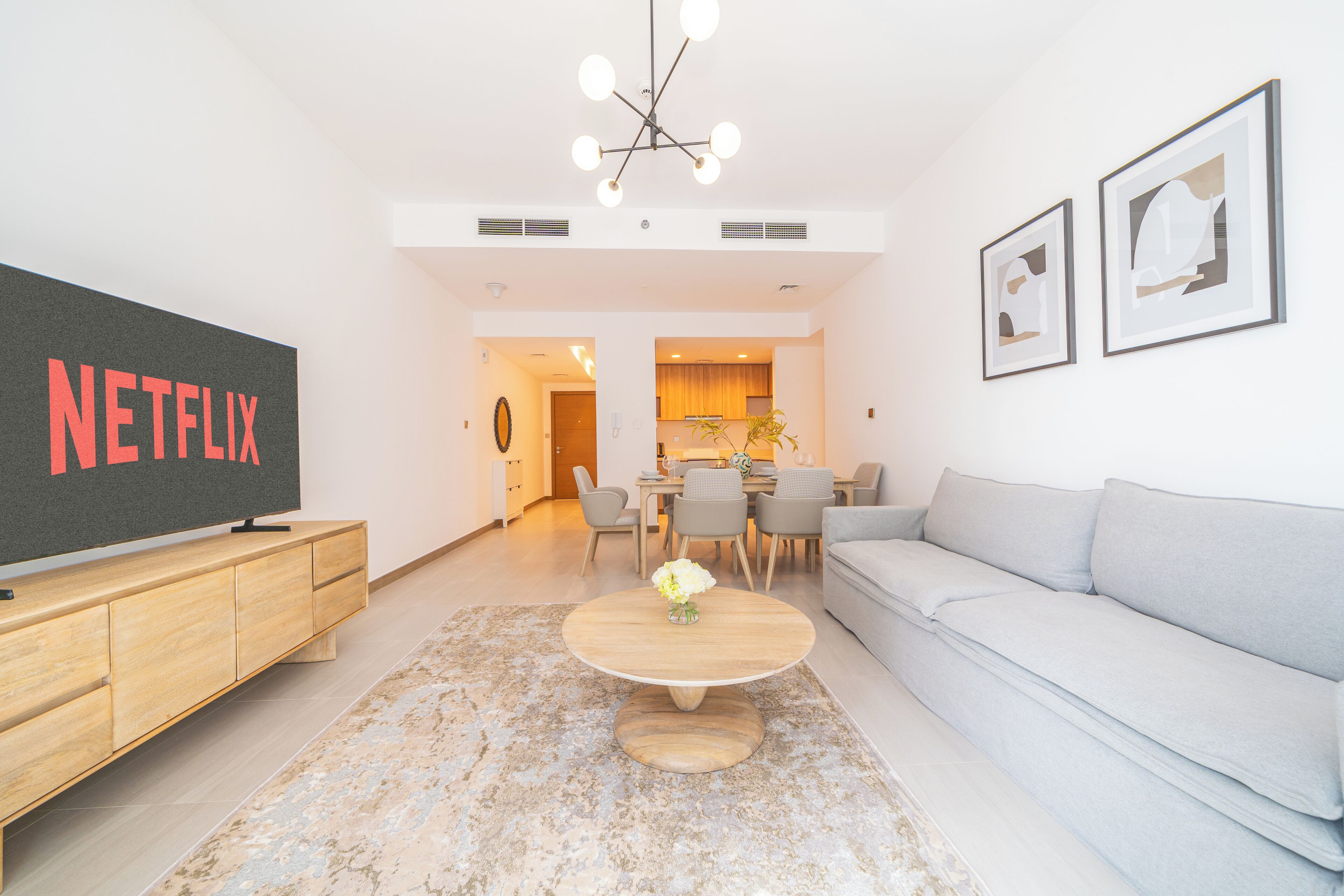 A modern living room with a flat-screen TV, a grey sofa, a wooden coffee table, and a dining area in the background.