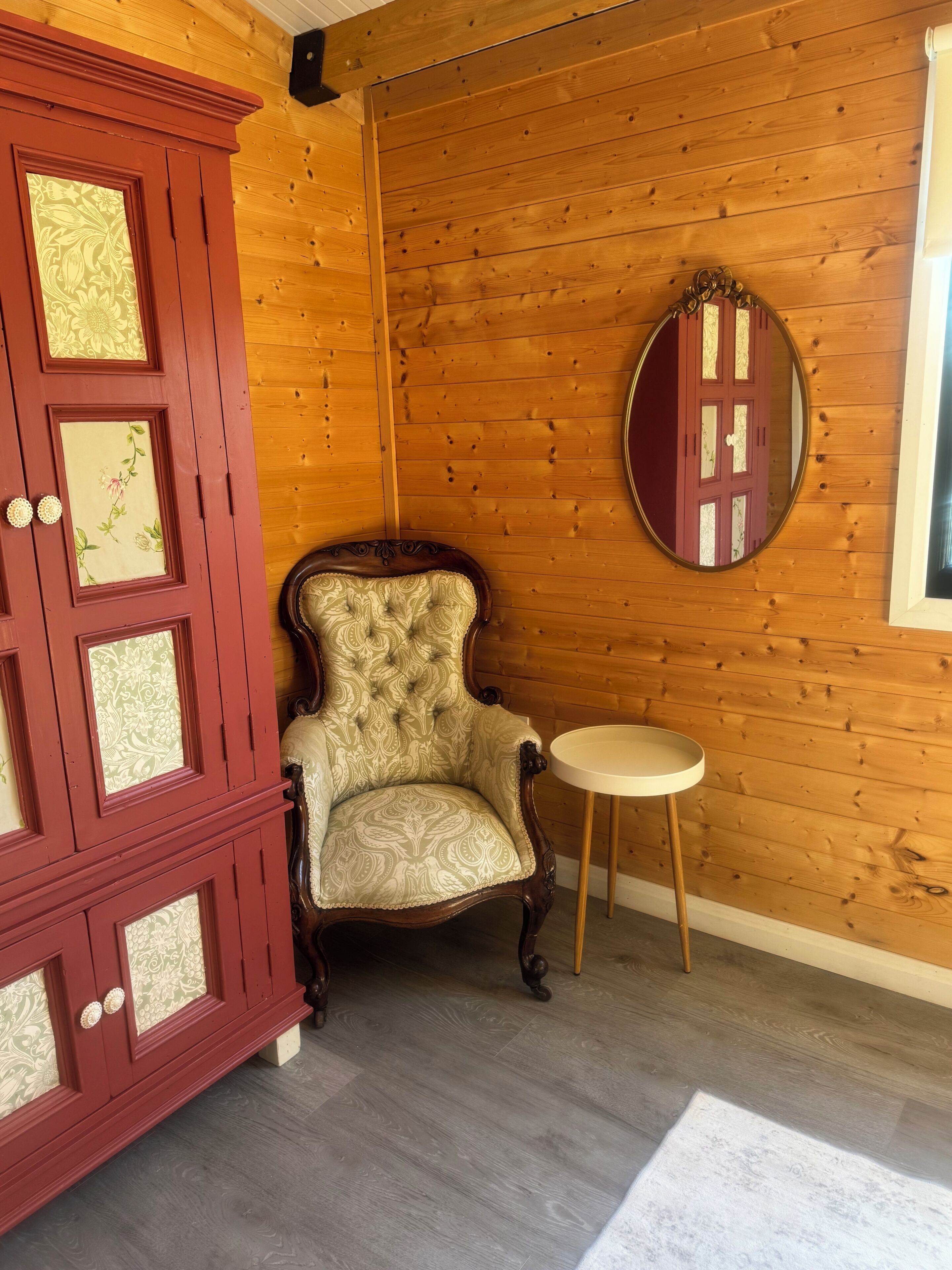 A room with a wooden paneled wall, a red cabinet with decorative glass panels, a patterned armchair, a round mirror, and a small round table.