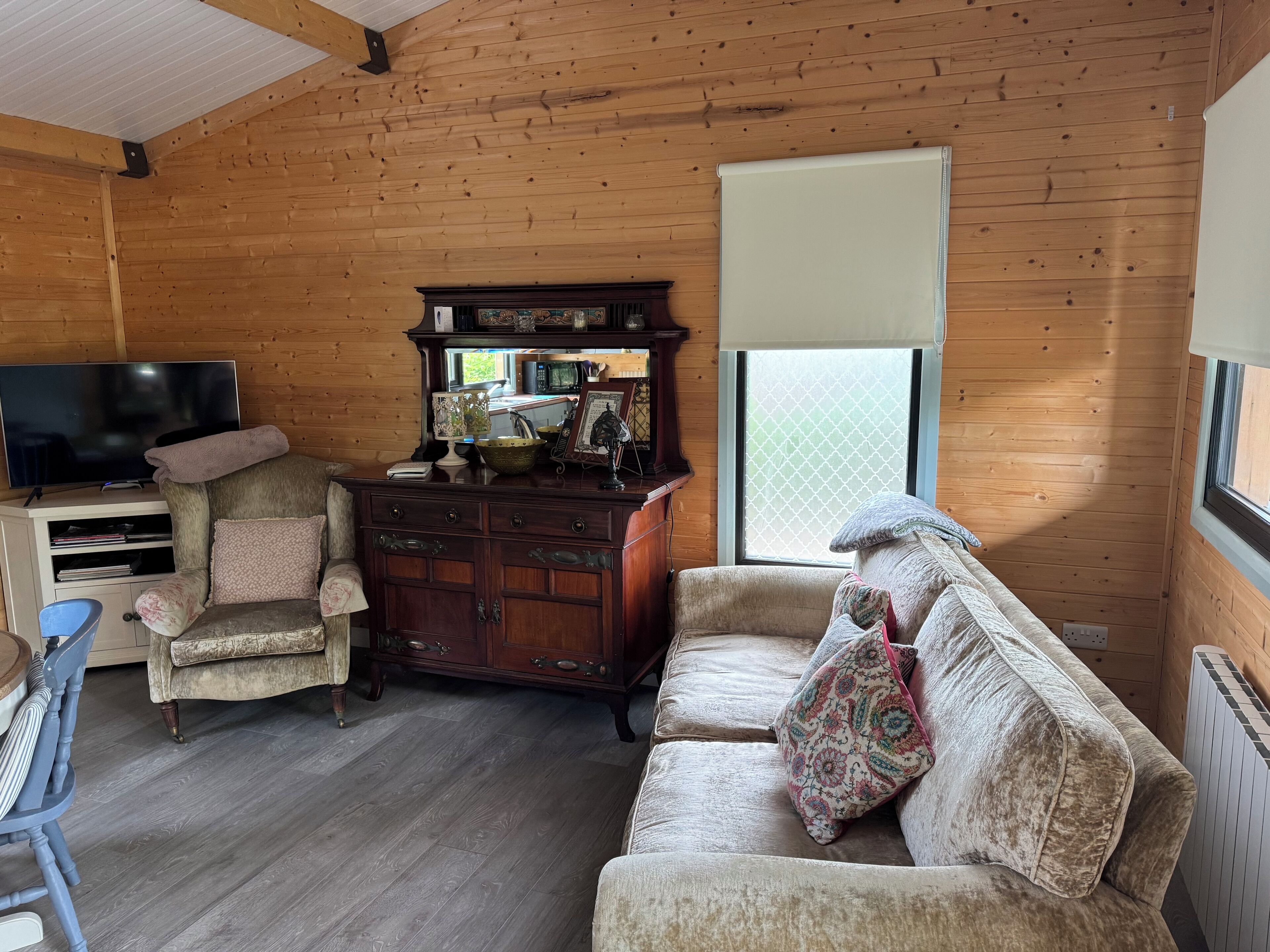 L’intérieur d’un chalet accueillant, avec un plafond en bois, un téléviseur posé sur un meuble en bois, une commode ancienne, un fauteuil rembourré et un canapé agrémenté de coussins à motifs.