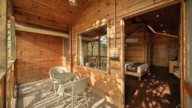 A wooden cabin with a balcony, wicker furniture, and a bed. - Aavaas Hill Resort (Lansdowne)