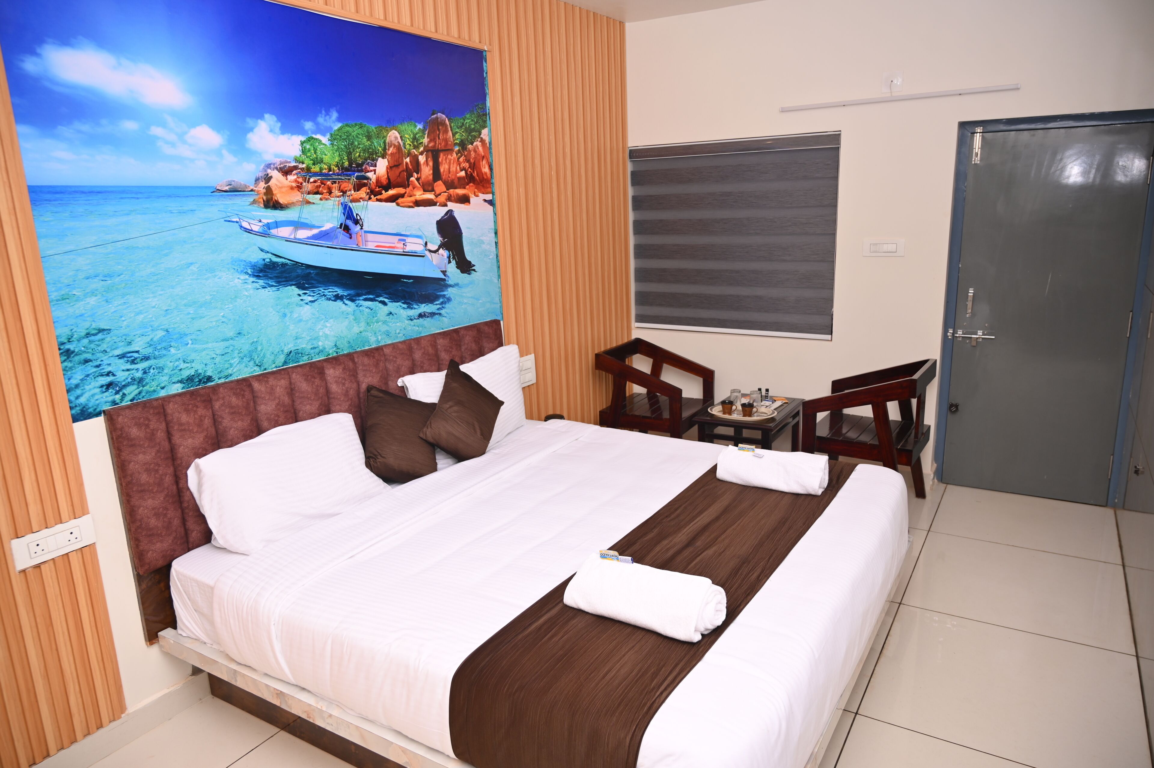 Ein Hotelzimmer mit einem großen Bett, einem Gemälde von einem Boot auf dem Wasser, einem hölzernen Kopfteil, einem kleinen Tisch mit einem Stuhl und einer Tür.