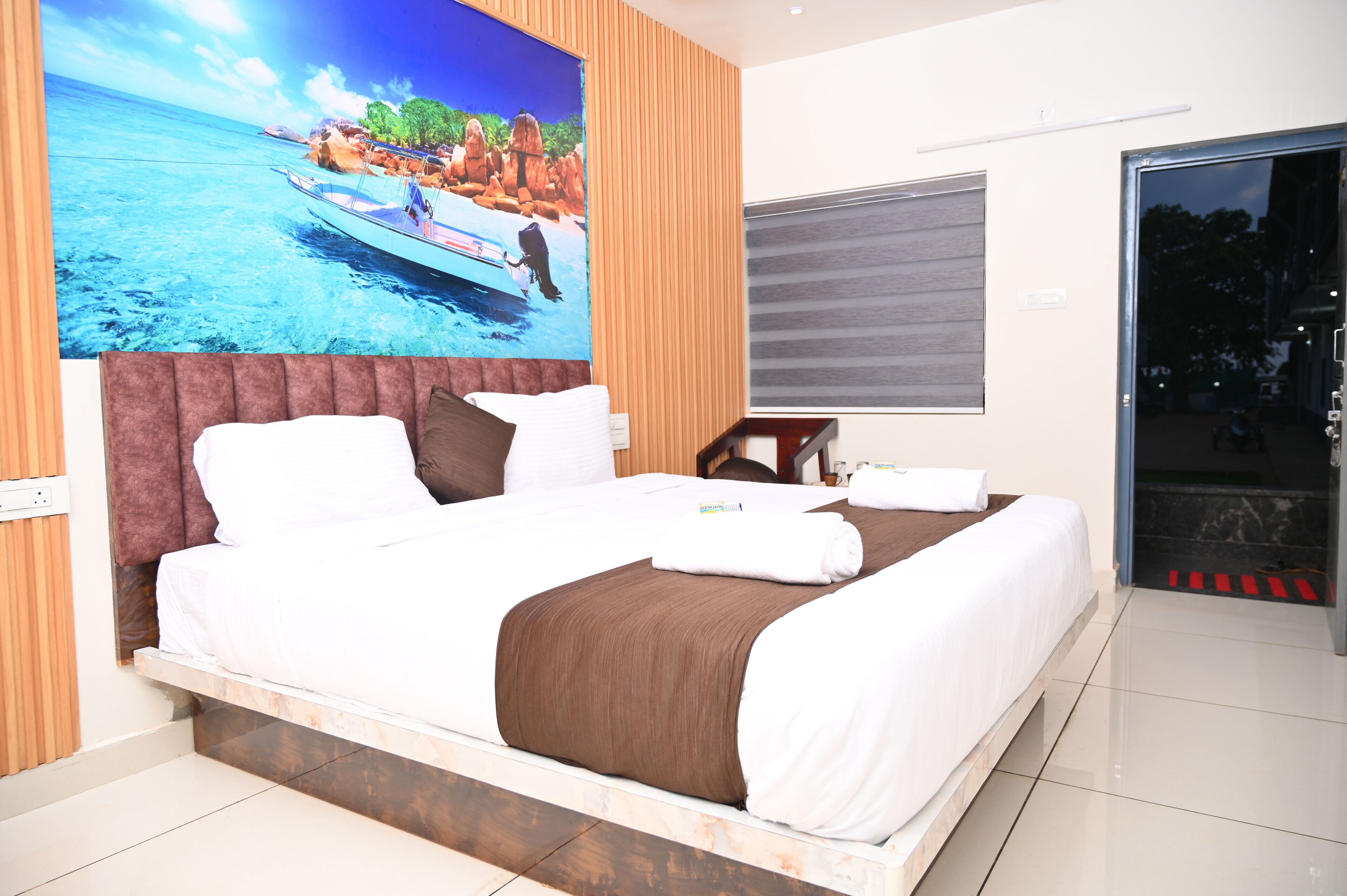 Habitación de hotel con una cama grande, un cuadro de una playa, una ventana con vistas y una puerta que da a un balcón.