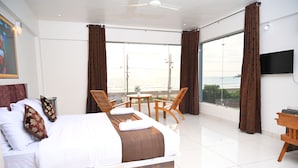 Une chambre d’hôtel avec un grand lit, un balcon avec une table et des chaises, une télévision et une vue sur la mer.