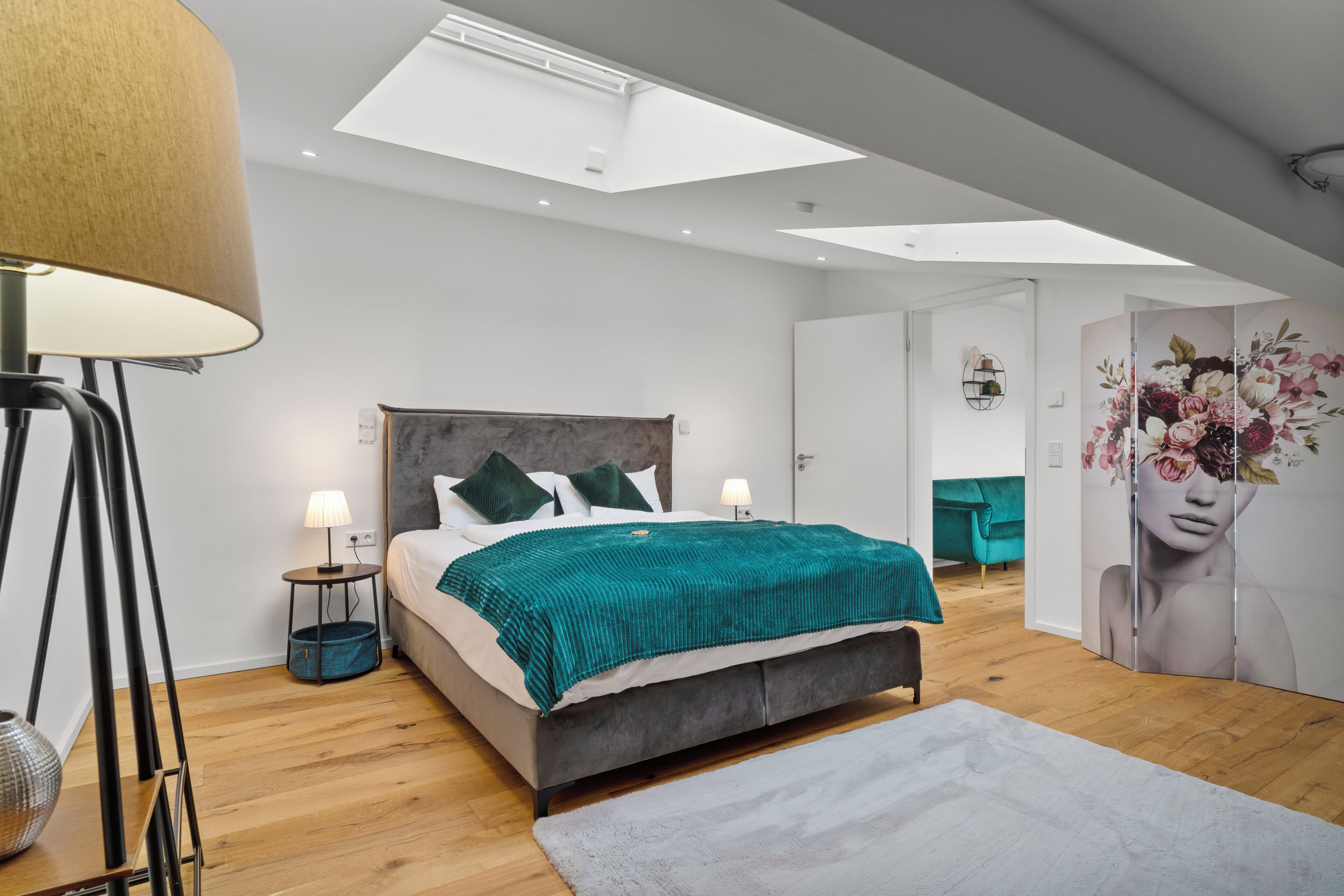 A modern bedroom with a skylight, a bed with a teal blanket, a side table with a lamp, and a large painting on the wall.