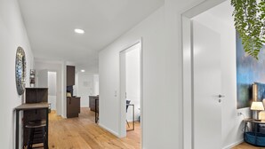 A hallway with wooden flooring, white walls, and a door leading to a room with a blue chair and a lamp. - Lit Living-Luxury-Box Spring - Parking (Hemsbach)