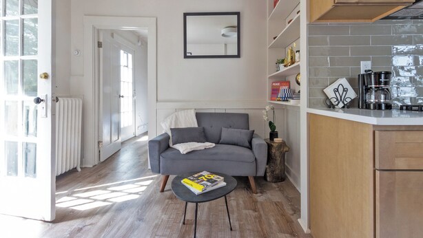 A modern living room with a grey sofa, a coffee table with a magazine, and a kitchen area with a coffee machine and shelves. - Colorful Modern Hideaway In Downtown Woodstock - AC & WiFi (Woodstock)