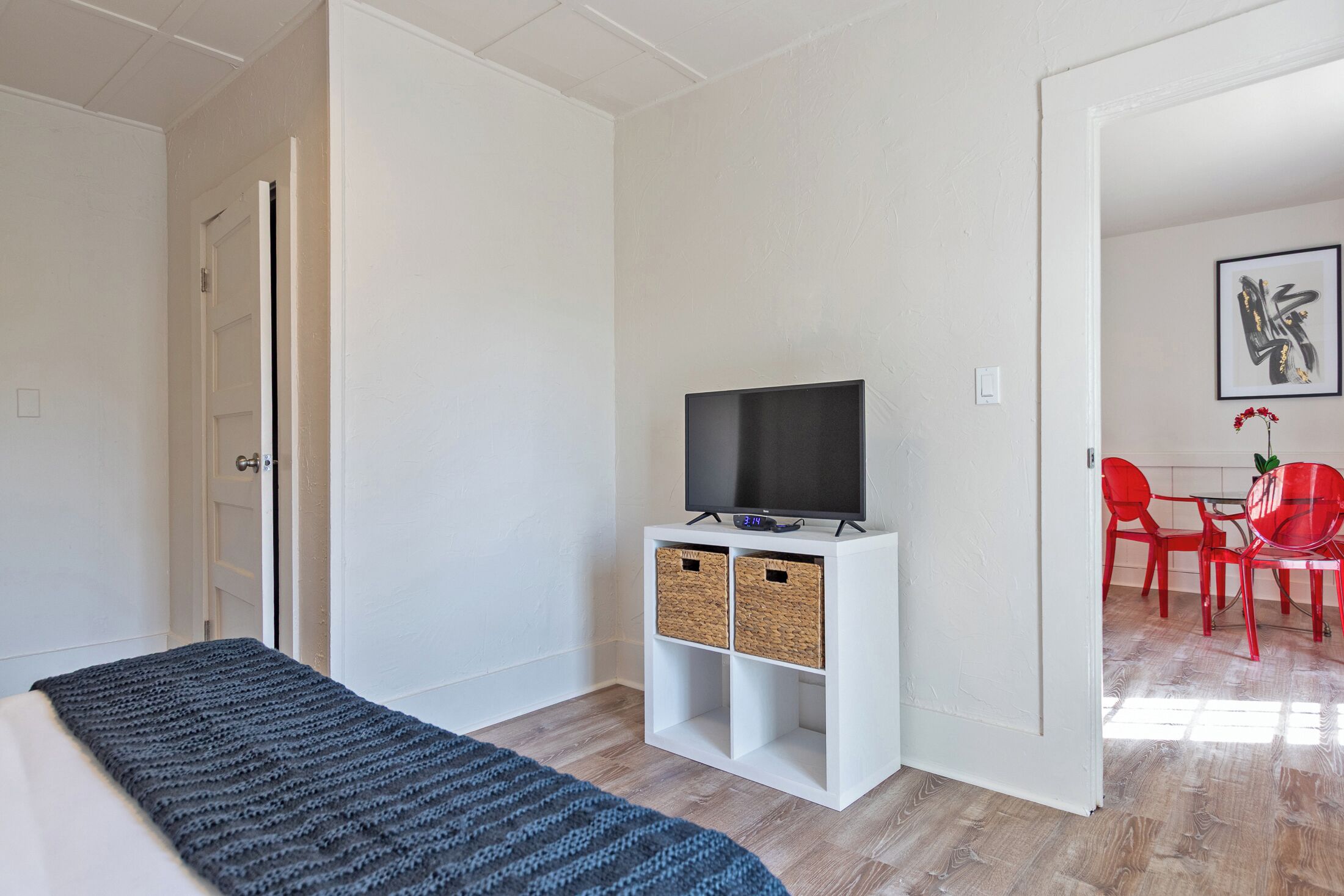 Una habitación con cama, un televisor sobre un soporte blanco, una silla roja y una puerta que da a otra habitación.