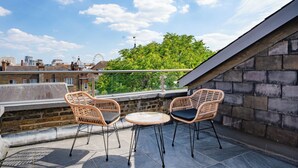 Una terraza en la azotea con dos sillas de mimbre y una pequeña mesa redonda, con vistas a un paisaje urbano y una noria.