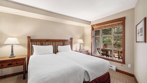 A bedroom with a large bed, two bedside tables with lamps, a wooden headboard, a window with a view of trees, and a framed picture on the wall. - Whistler - Horstman House - 2 Bedroom #403 (Whistler)