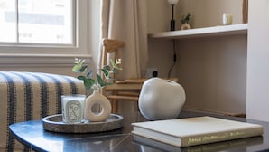 A neatly arranged room with a striped sofa, a small round table with a book, a candle, and a vase with a plant. - Be London - St John's Wood Residences (London)