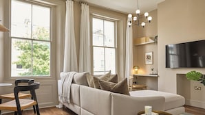 A living room with a sofa, two chairs, a TV, and a window with curtains. - Be London - St John's Wood Residences (London)