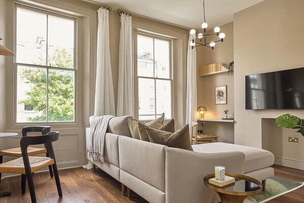 A living room with a sofa, two chairs, a TV, and a window with curtains. - Be London - St John's Wood Residences (London)