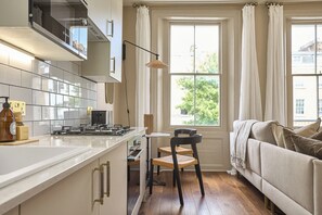 A modern kitchen with white cabinets, a stove, and a dining area with a chair and table. - Be London - St John's Wood Residences (London)