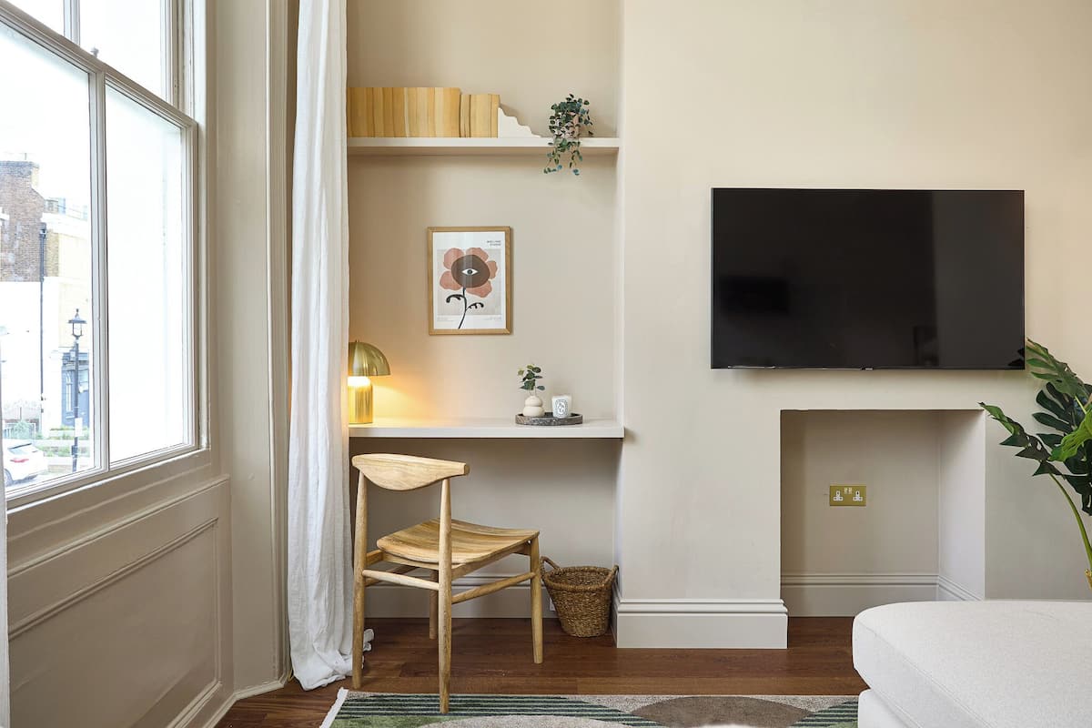 Een kamer met een houten stoel, een bureau met een lamp, een wandgemonteerde televisie en een raam met gordijnen.