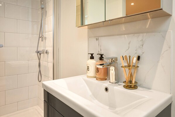 A modern bathroom with a glass-enclosed shower, a white sink with marble backsplash, and toiletries.