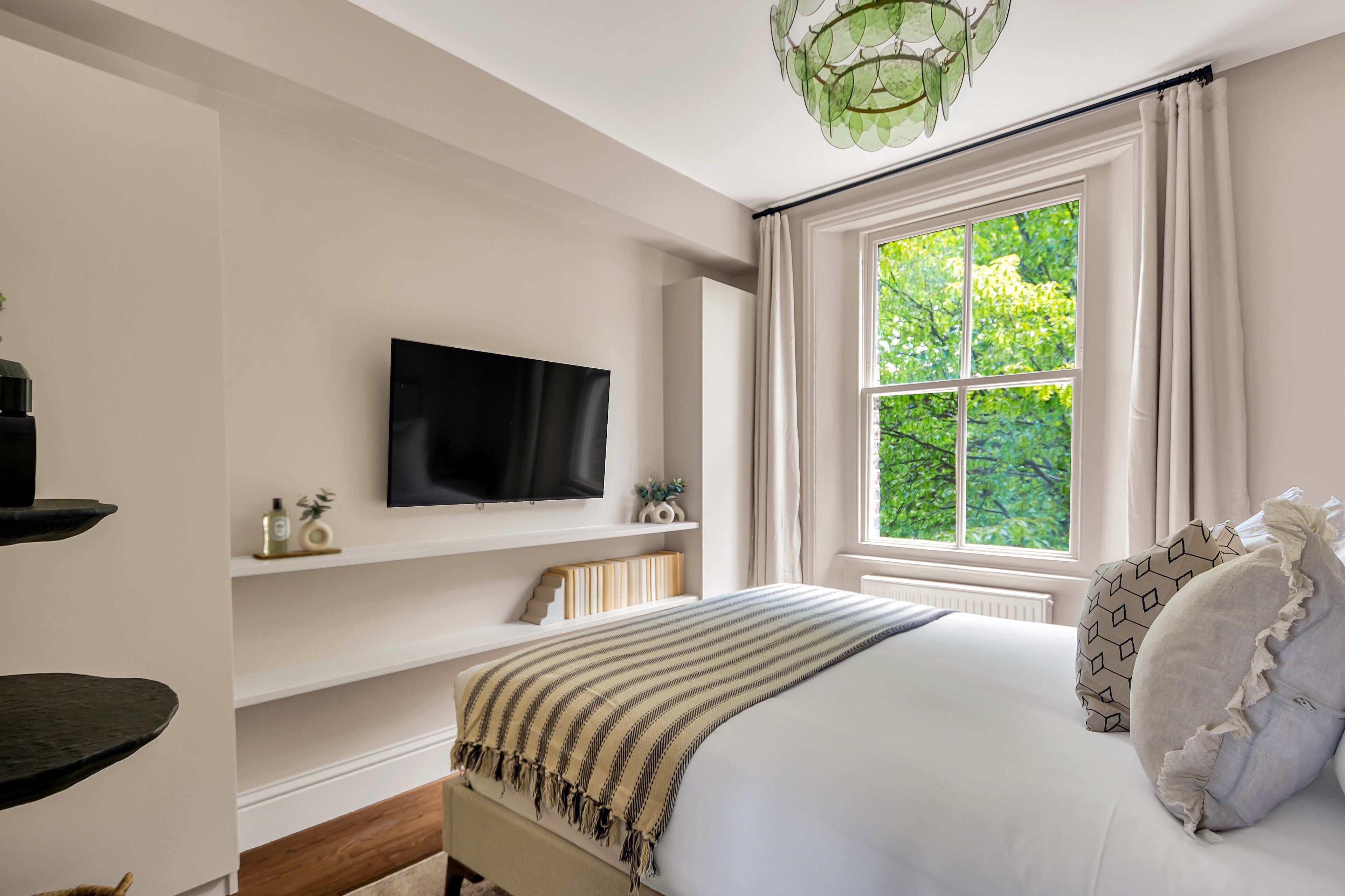 Un dormitorio con cama, televisor en la pared, ventana con cortinas y estante con adornos.