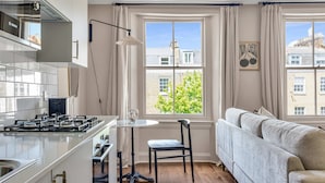 A modern kitchen with a gas stove, a dining table, and a chair. There is a window with curtains, a sofa, and a framed picture on the wall. - Be London - St John's Wood Residences (London)