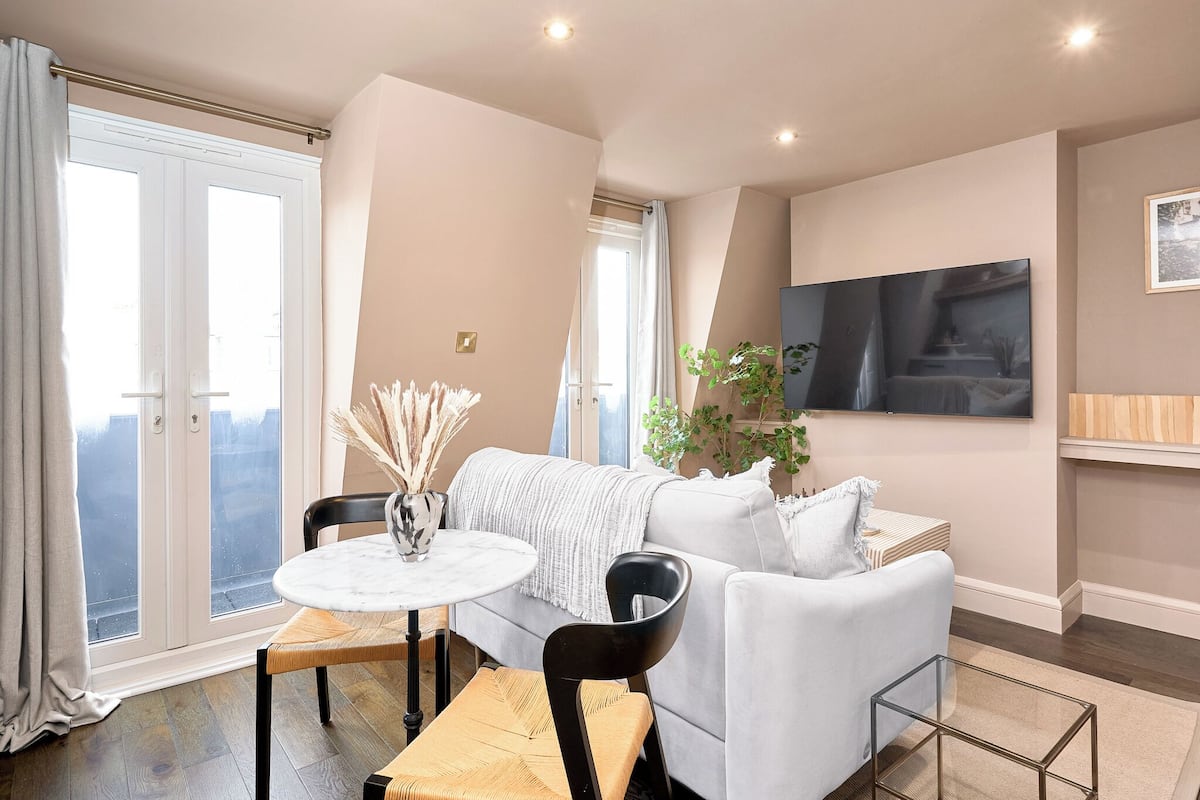 Een moderne woonkamer met een witte bank, een ronde tafel en een zwarte stoel. Er is een groot raam met gordijnen, een flatscreen-tv aan de muur en een potplant.