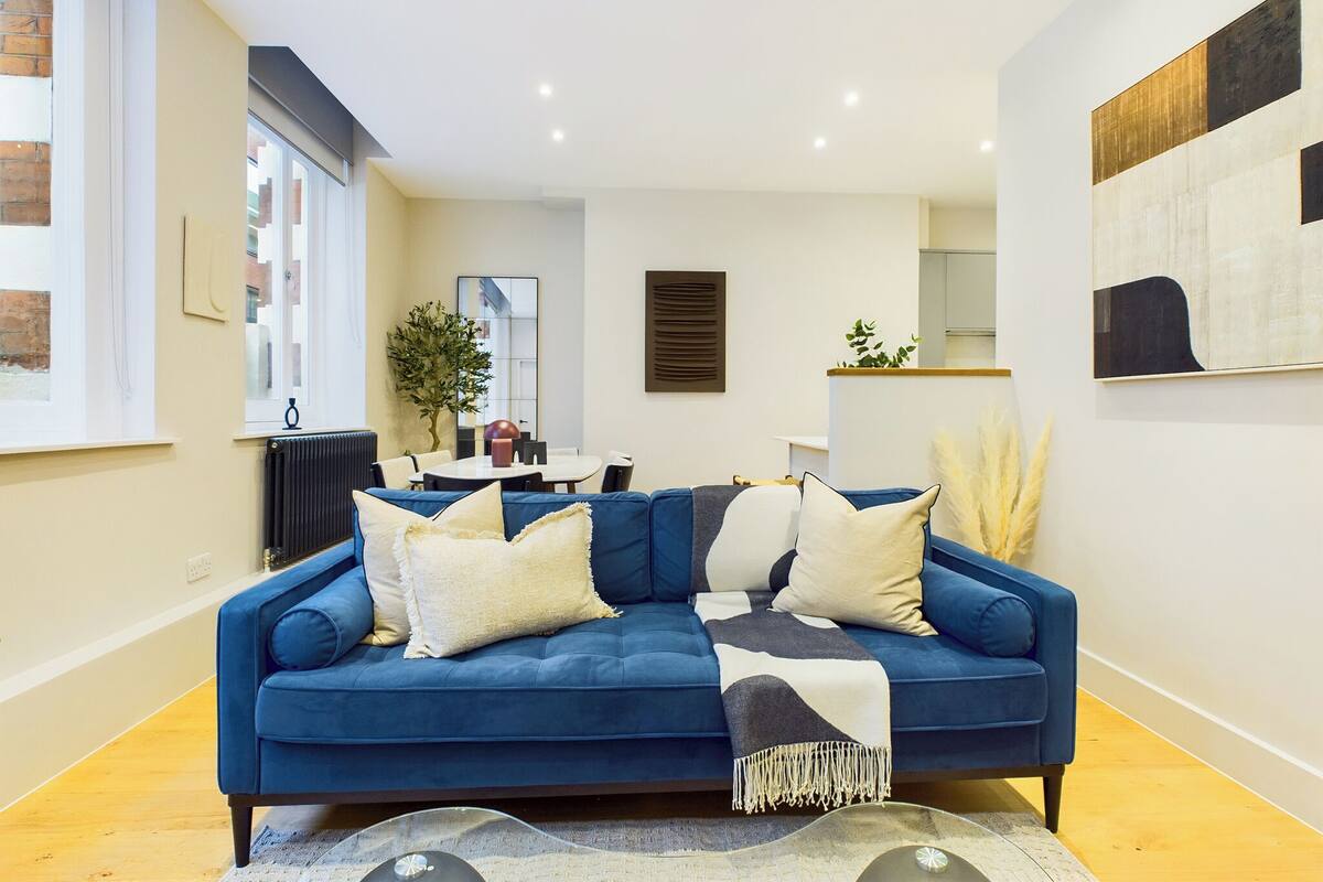 A modern living room with a blue sofa, a dining table, and a kitchen area in the background.