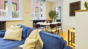 A modern living room with a blue sofa, a dining table, and chairs. There are windows with a view of buildings outside. - Be London - Piccadilly Circus Residences (London)