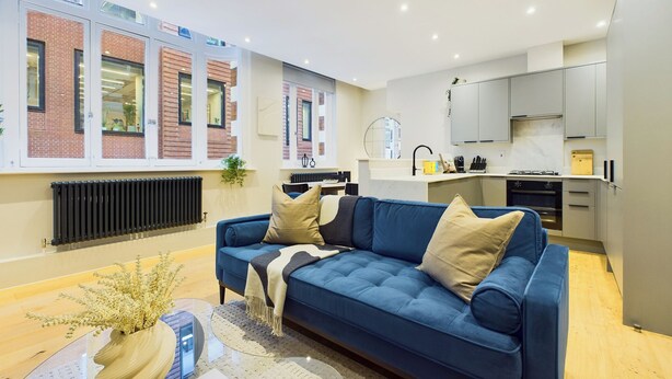 A modern apartment with a blue sofa, a kitchen with grey cabinets, and a view of a brick building through large windows. - Be London - Piccadilly Circus Residences (London)