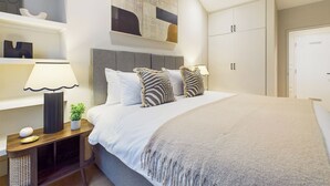 A modern bedroom with a large bed, a bedside table with a lamp, a small plant, and a wall art piece above the bed. - Be London - Piccadilly Circus Residences (London)