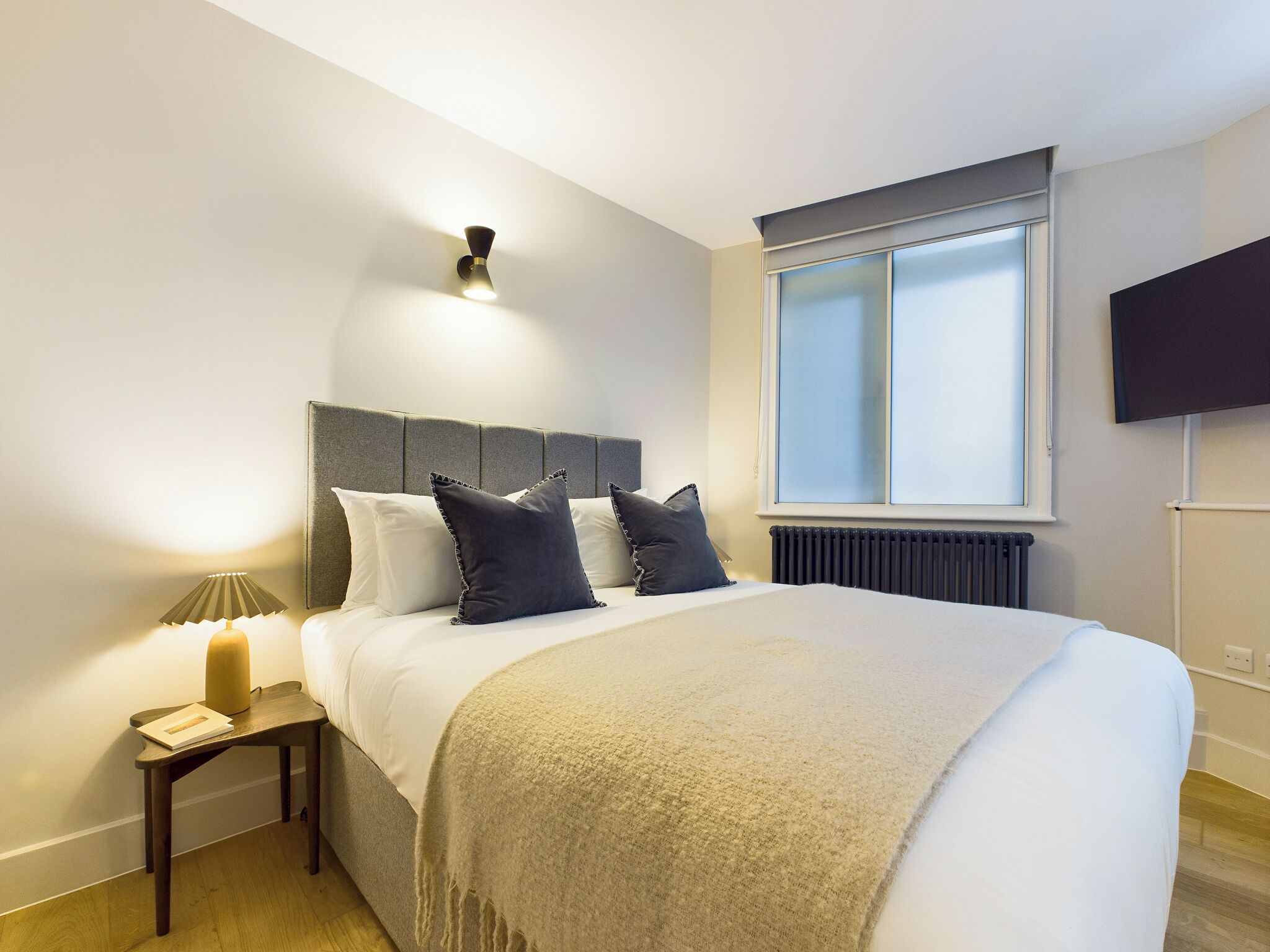 A modern bedroom with a large bed, a bedside table with a lamp, a window with blinds, and a mounted TV.