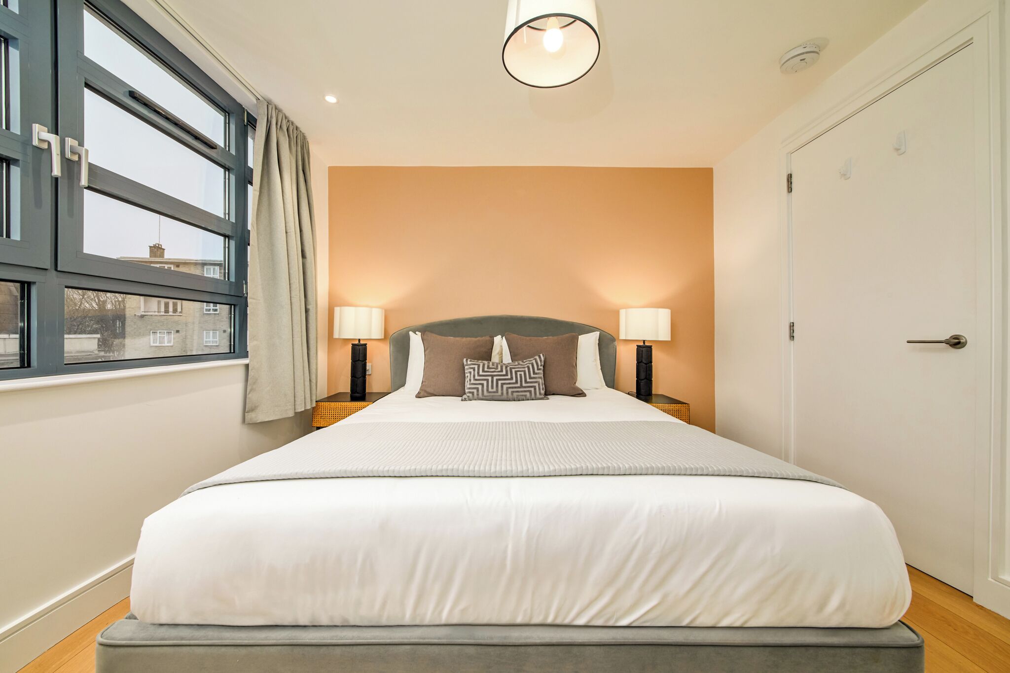 A neatly made bed with a headboard, two bedside lamps, and a window with curtains.