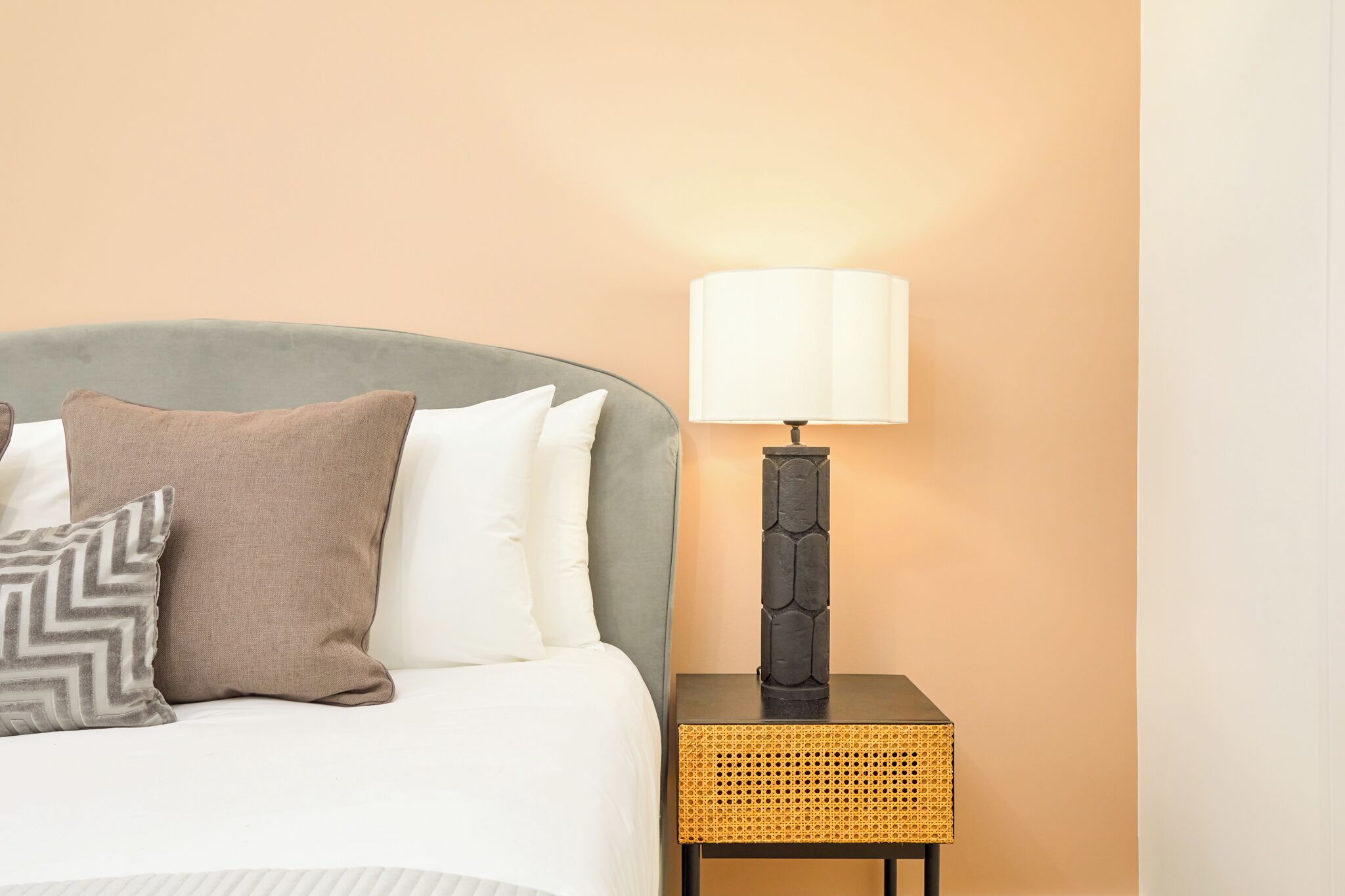 A neatly made bed with a grey headboard, a bedside lamp, and a woven nightstand.