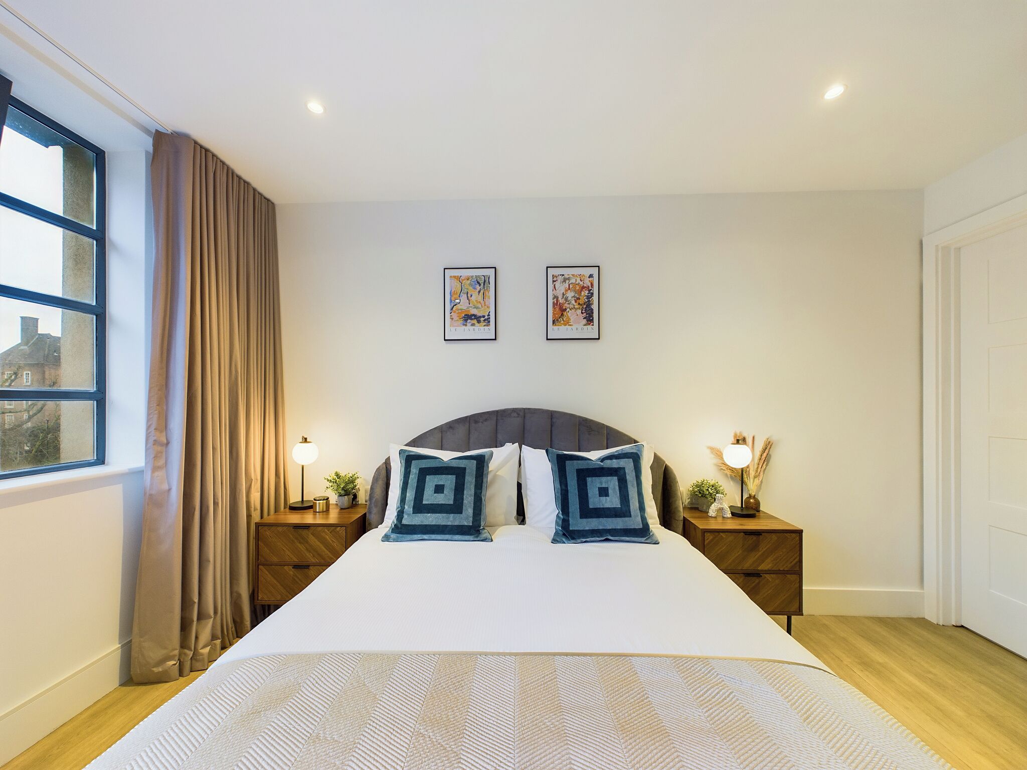 A neatly arranged bedroom with a large bed, two bedside tables with lamps, a window with curtains, and two framed pictures on the wall.