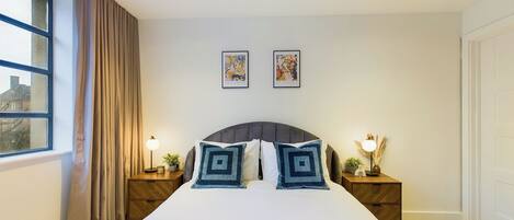 A neatly arranged bedroom with a large bed, two bedside tables with lamps, a window with curtains, and two framed pictures on the wall.