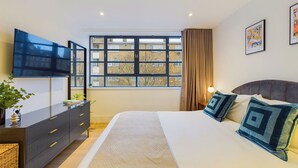 Una camera da letto moderna con un letto grande, una TV a schermo piatto montata a parete, una finestra vista edifici e un comodino con lampada.