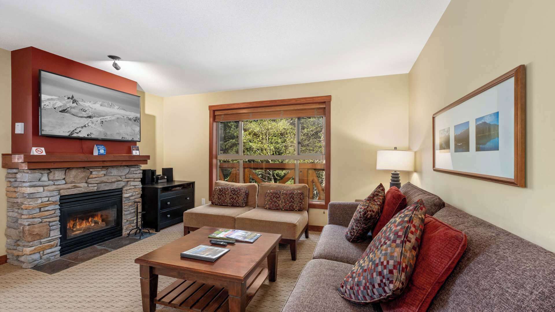 A cozy living room with a stone fireplace, a large window with a view of greenery, a sofa with patterned cushions, and a coffee table.