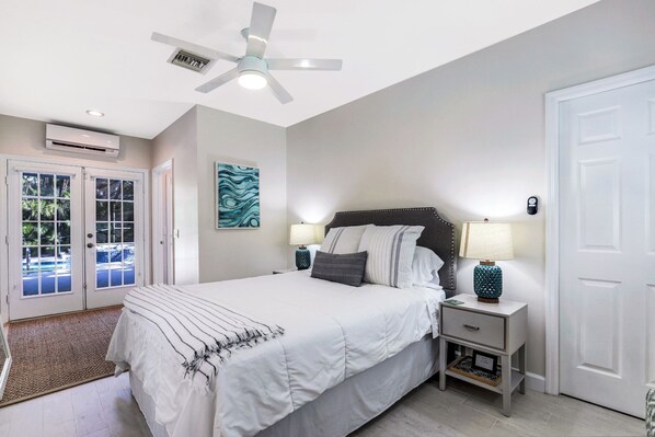 A bedroom with a bed, two bedside tables with lamps, a ceiling fan, an air conditioning unit, and a door with a view of the outside.