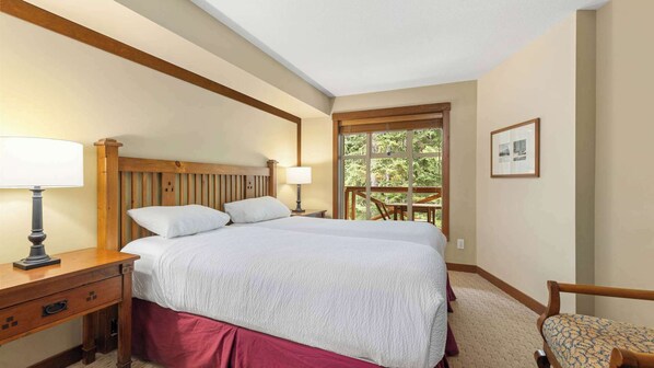 Un dormitorio con cabecera de madera, una cama grande con ropa blanca, una mesita de noche con lámpara, una silla y una ventana con vista a zonas verdes.