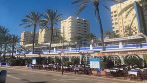 A coastal urban area with palm trees, a restaurant named "PIZZA PASTA CARNE PESCADO," and high-rise buildings in the background. - Estepona Marina Apartment, Costa Del Sol (Estepona)