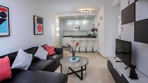 A modern living room with a black leather sofa, a round coffee table, and a TV on a stand. A kitchen is visible in the background with white cabinets and a bar stool. - Estepona Marina Apartment, Costa Del Sol (Estepona)