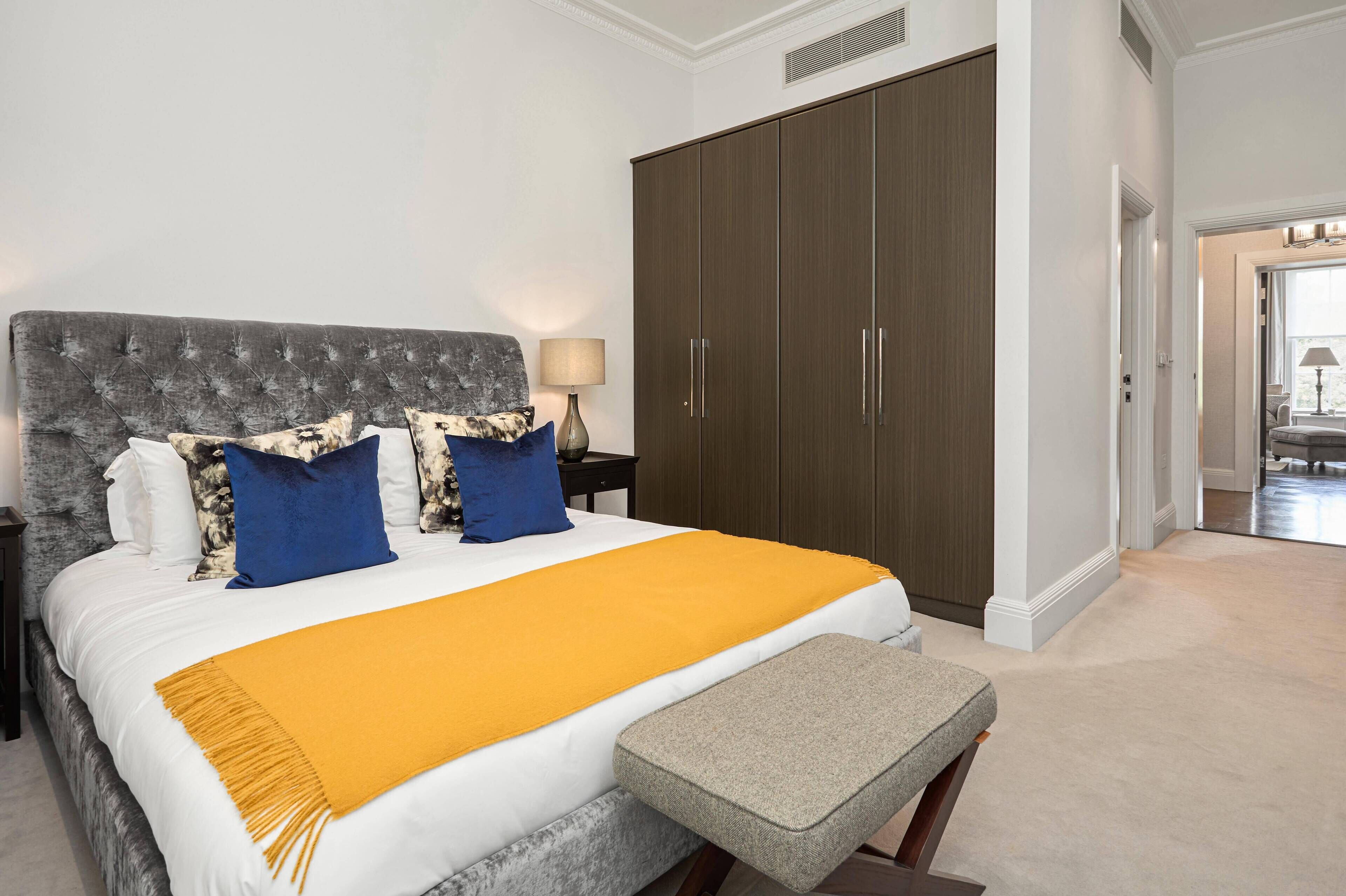 A bedroom with a large bed, a grey upholstered headboard, a yellow throw blanket, and a grey bench.