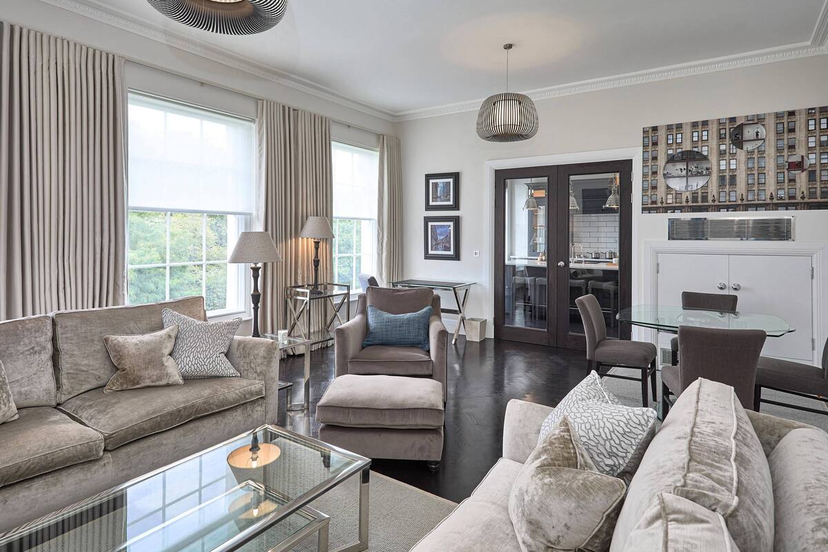 A spacious living room with a large window, a glass coffee table, and a modern chandelier.