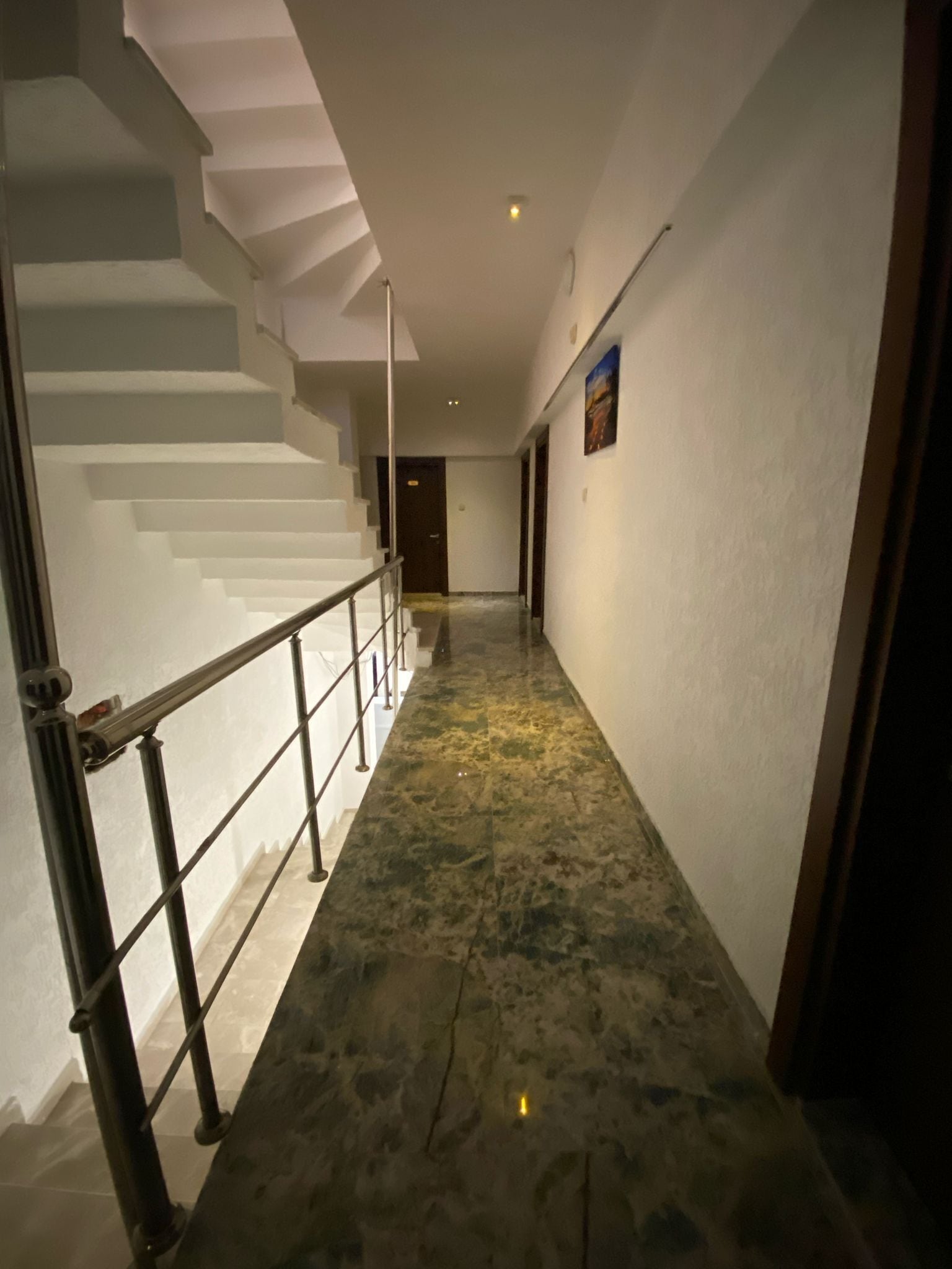 A hallway with a marble floor, a staircase with a metal railing, and a door at the end.
