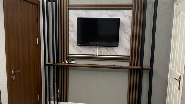 A modern bedroom with a large TV mounted on the wall, a wooden wardrobe, a bedside table with a towel and a book, and a blue mat indicating the bathroom. - Deniz Butik Hotel (Tekirdag)