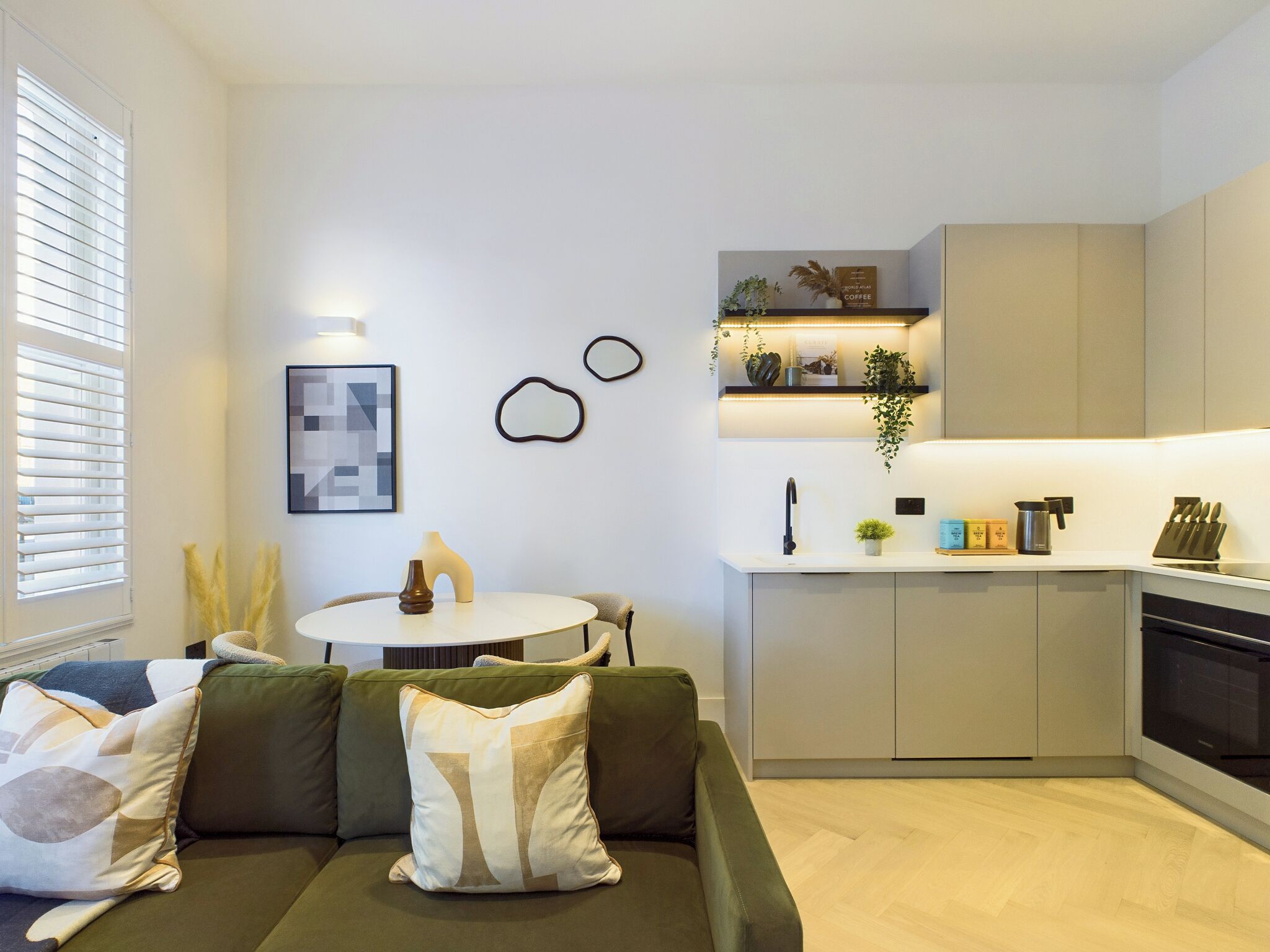 A modern kitchen with a white countertop, built-in cabinets, and a black oven. There is a dining area with a round table and chairs, and a green sofa with decorative pillows.