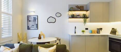 A modern kitchen with a white countertop, built-in cabinets, and a black oven. There is a dining area with a round table and chairs, and a green sofa with decorative pillows.