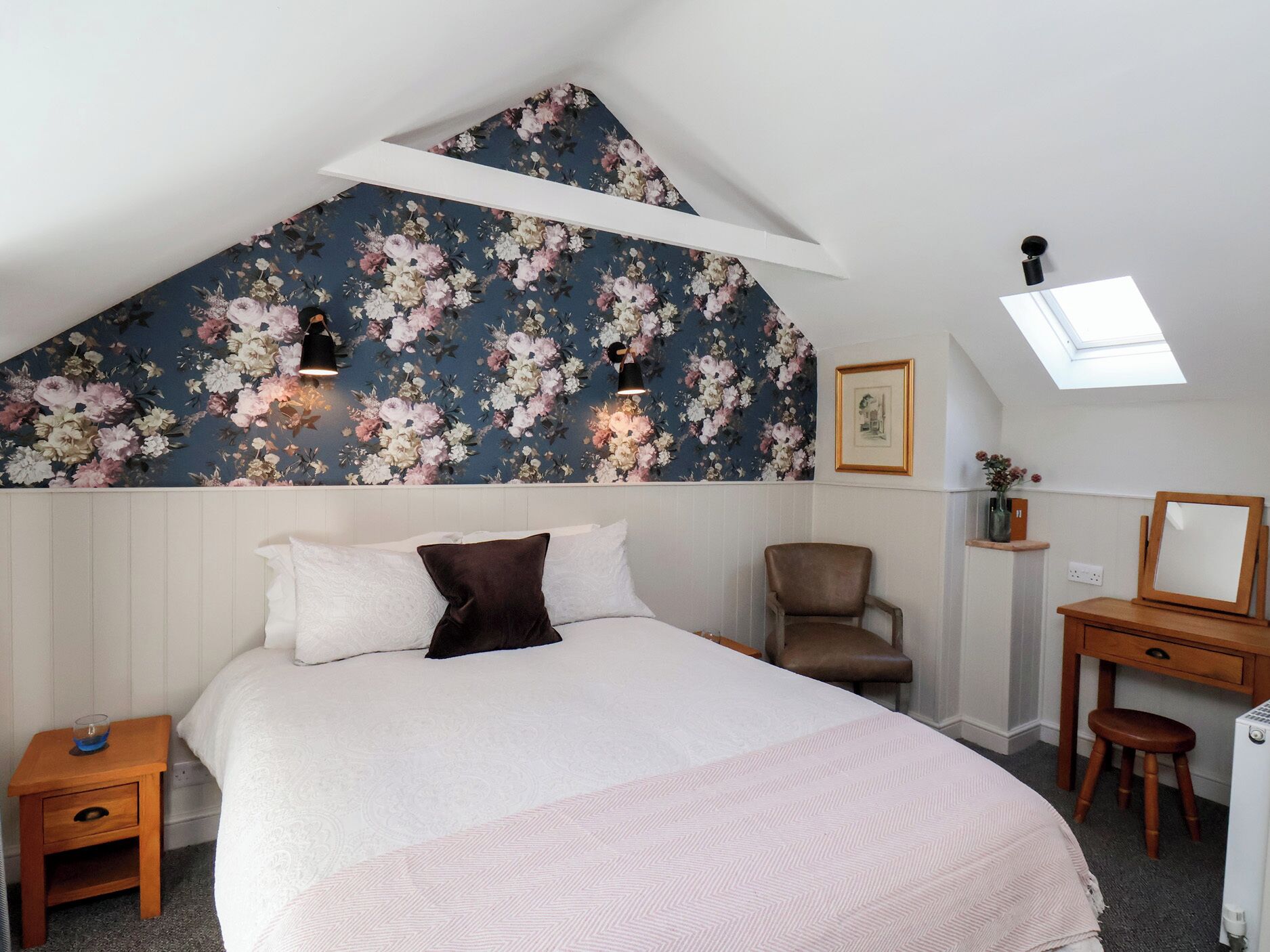 Un dormitorio con techo inclinado, papel tapiz floral, cama con ropa de cama blanca y marrón, una silla, un escritorio con espejo y un tragaluz.