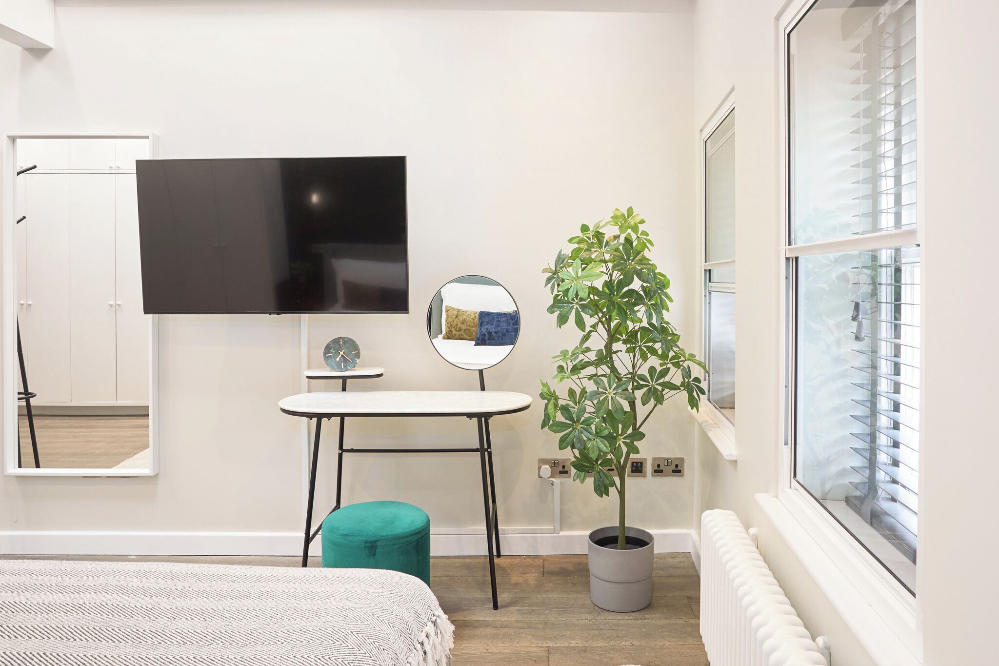A modern bedroom with a bed, a TV, a small table, a mirror, a potted plant, and a window with blinds.
