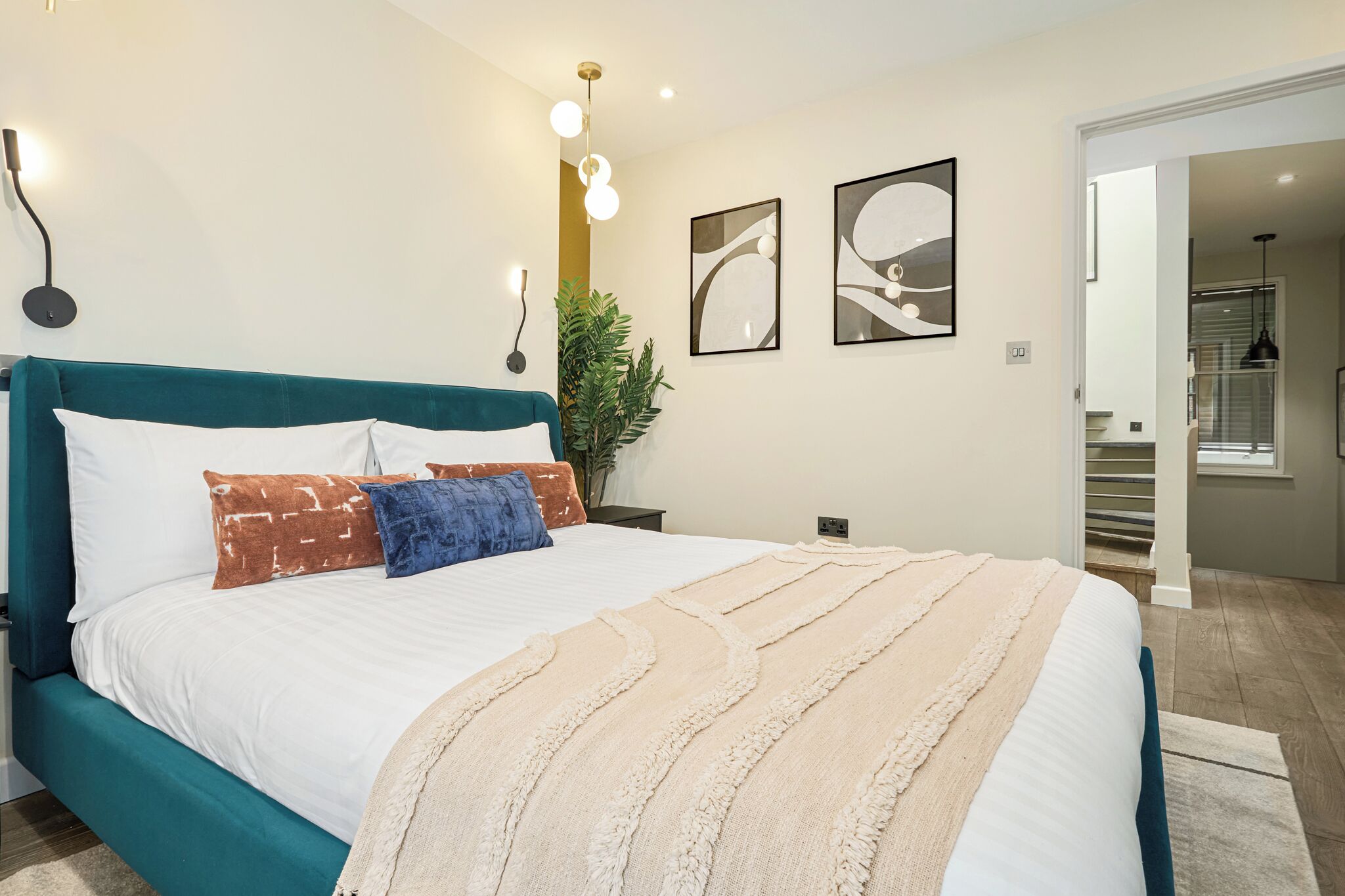 A modern bedroom with a teal headboard, white bedding, and a beige throw blanket. There are abstract art pieces on the wall and a potted plant beside the bed.