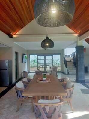 A spacious dining area with a large wooden table and wicker chairs, a hanging light fixture, and a view of the outdoors through large windows.