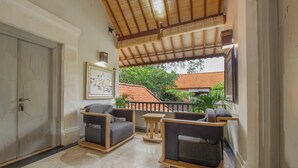 A balcony with seating, a door, and a view of a building with a red roof. - Dampati Villas (Denpasar)