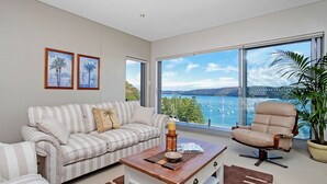 A modern living room with a striped sofa, a single armchair, a wooden coffee table, and a large window offering a view of a lake and boats. - The Commodore Penthouse (Palm Beach)